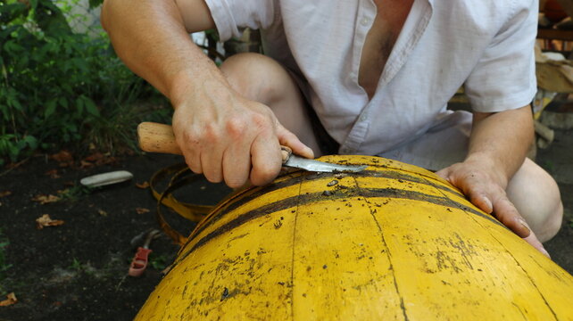 A man scrapes dirt off the surface of an old yellow wooden barrel with a knife. The repairs are taking place on the ground, with removed hoops, tools, and green foliage visible in the background.