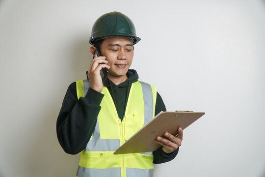 Focused Engineer in Safety Vest Talking on Phone and Reviewing Clipboard