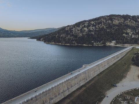 Aerial view of Lac des Bouillouses dam with a long stone wall, calm blue water, surrounding pine forests, and mountains in Pyrenees-Orientales, Occitanie, France.