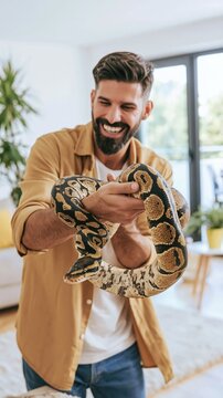 Smiling man happily holding a ball python in a modern living room, caring for an exotic pet