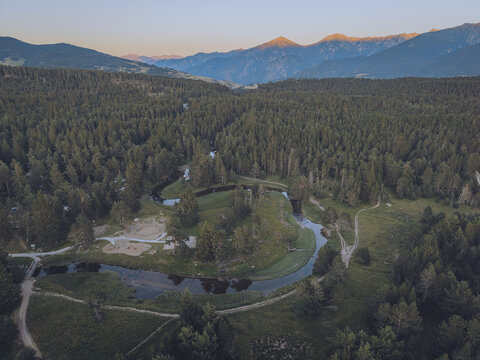 Aerial view of Le Petit Canada featuring a winding river flowing through a dense pine forest with mountain peaks in the background in Bolquere, Occitanie, France.