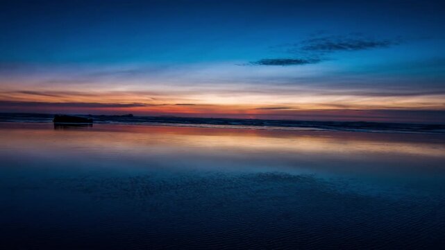 Colorful orange and blue twilight sky reflects onto wet sand as ocean waves gently wash ashore along a tranquil beach wide shot