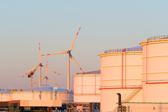 Large oil storage tanks at an industrial refinery facility with wind turbines in the background. Petroleum fuel storage tanks near a green energy wind farm.