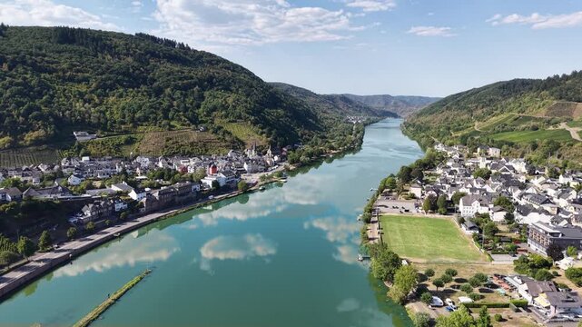 Aerial drone view of Bullay on the Moselle River in Rhineland-Palatinate, Germany, surrounded by vineyards and forested hills. Scenic Moselle Valley landscape with reflections of clouds in the calm ri