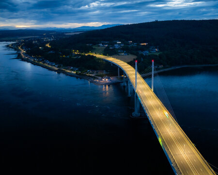 Aerial view of Kessock Bridge illuminated at night over the Beauly Firth with surrounding coastal landscape and city lights in Inverness, Scotland, United Kingdom.