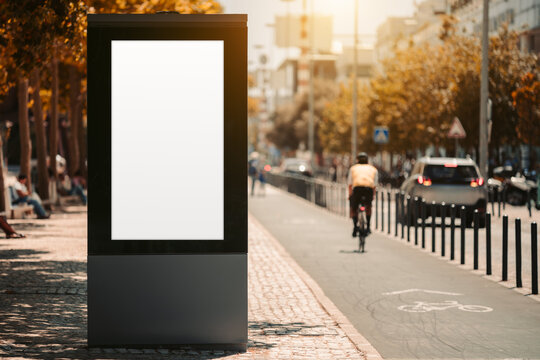 Blank vertical billboard mockup dominates the foreground beside a sunlit city bike lane, with cyclist silhouette, traffic and autumn trees softly blurred to the right on a copy space area