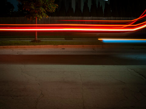 Long exposure night photography showing vibrant red and blue light trails from moving vehicles speeding past a suburban street and trees