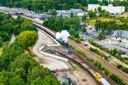 Aerial view of a steam train arriving at Aviemore railway station surrounded by lush green trees and buildings in Aviemore, Scotland, United Kingdom.