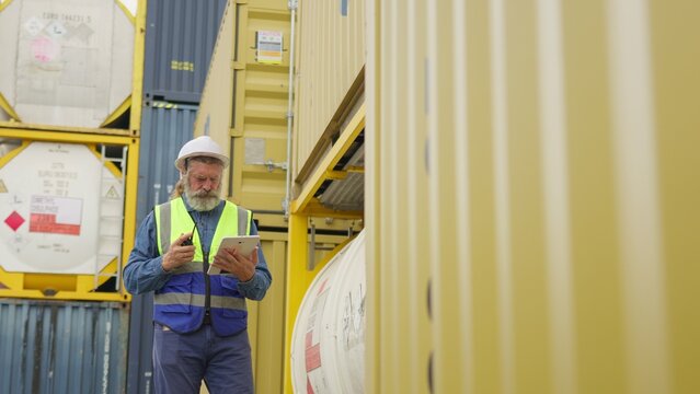 A mature foreman uses a digital tablet for inventory management, inspecting cargo containers at an industrial shipping port to ensure supply chain efficiency.
