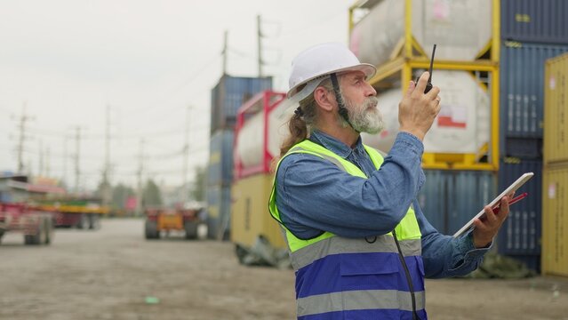 An experienced foreman supervises cargo operations with a radio and tablet at a bustling shipping yard, coordinating logistics for supply chain management.