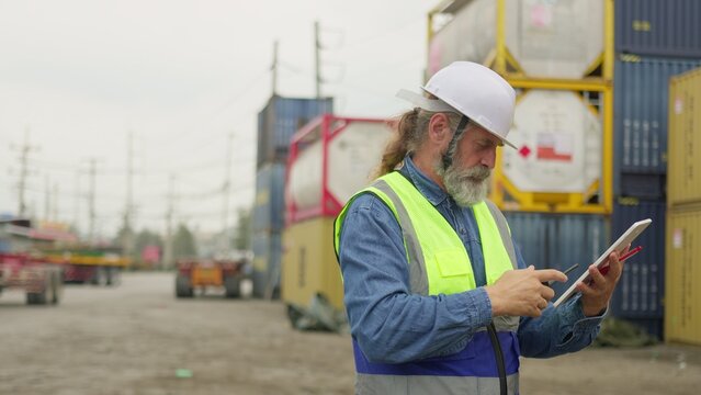 An experienced foreman supervises cargo operations with a radio and tablet at a bustling shipping yard, coordinating logistics for supply chain management.
