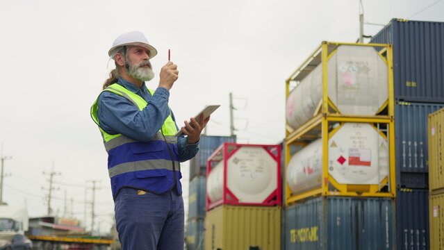 A senior logistics manager in a hard hat uses a digital tablet for inventory control while inspecting cargo at an industrial shipping container terminal.