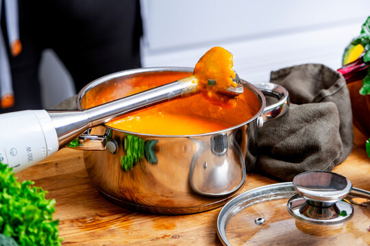 Woman makes pureed pumpkin soup with hand blender in metal pot. Autumn seasonal hot comfort food concept.
