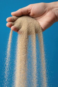 Close up of human hand releasing fine sand. Particles flow through fingers against blue background. Concept of time passing, lost wealth, hourglass motion. Sand pours down in slow motion stream for