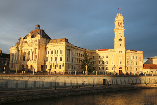 Historic City Hall Palace of Oradea, Romania, illuminated by warm evening sunlight beside the Crișul Repede River under a dramatic dark sky.