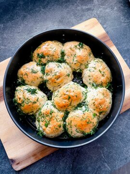 Freshly baked round bread , pampushki, garlic rolls topped with herbs arranged in a black bowl on a wooden cutting board against a dark stone surface