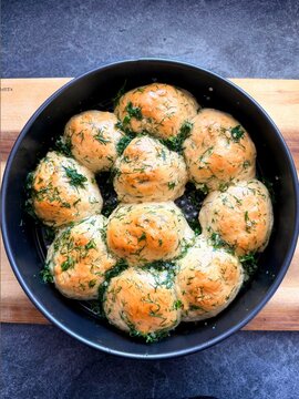 Freshly baked round bread , pampushki, garlic rolls topped with herbs arranged in a black bowl on a wooden cutting board against a dark stone surface