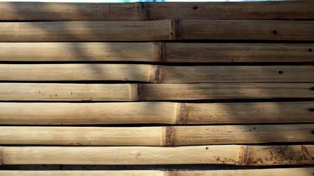 Close-up of wall made of natural bamboo logs arranged horizontally, showcasing texture, color variations, and cuts illuminated by sunlight