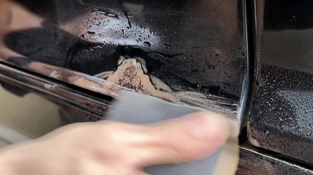 Close-up of a hand using sandpaper to remove rust from a car door trim, showing the gradual cleaning process with visible dirt and moisture on the surface