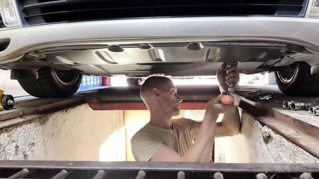 Male mechanic working under a car, using a wrench to tighten components, with tools and a workshop visible in the background, showcasing detailed automotive maintenance