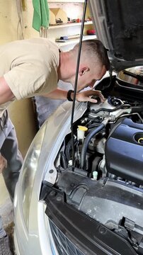 Male mechanic in beige shirt inspects car engine components under the hood in a garage, with tools and spare parts visible in the background