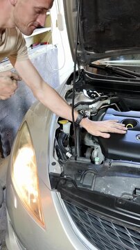 Male mechanic in beige shirt inspects car engine components under the hood in a garage, with tools and spare parts visible in the background