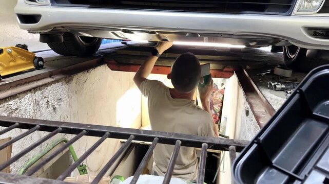 Male auto mechanic inspecting car components from beneath vehicle on a lift, showcasing detailed actions in a garage environment with tools and equipment visible
