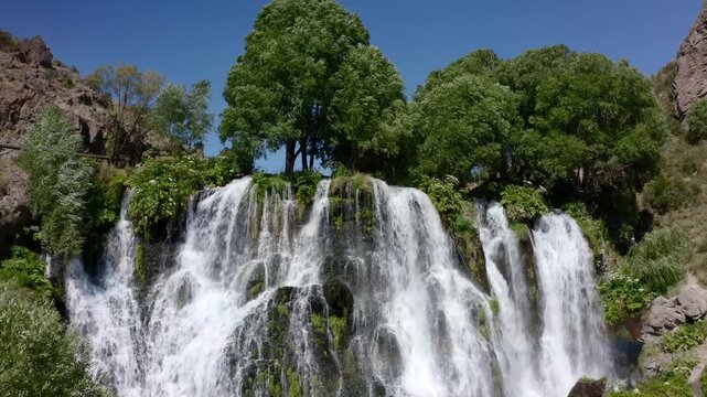 Scenic view of Shaki Waterfall cascading down basalt rocks framed by lush green trees and mountain landscape under blue sky in Syunik Province Armenia.