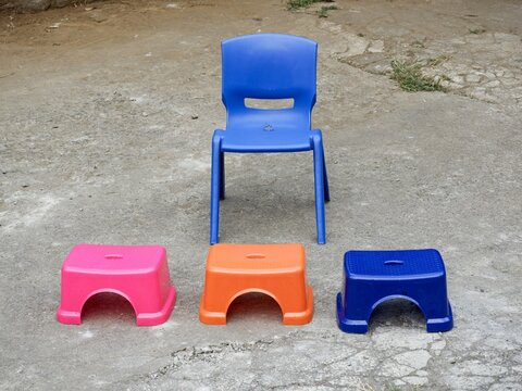 Bright blue plastic chair placed behind three small colorful stools in pink, orange, and blue on concrete ground.