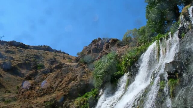 Close-up low angle shot of water drops spraying and splashing directly onto lens from powerful Shaki Waterfall cascades surrounded by green trees in Armenia.