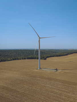 Aerial view of a single wind turbine standing in a golden wheat field with a dense forest in the background under a clear blue sky in Saint-Amand-sur-Fion, Grand Est, France.