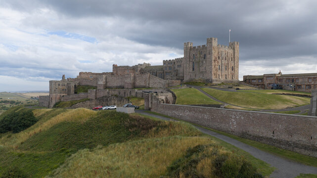Aerial view of Bamburgh Castle, a historic fortress with stone walls and towers, set on a grassy hill under a cloudy sky in Bamburgh, England, United Kingdom.