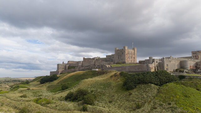 Aerial view of Bamburgh Castle perched on a rocky outcrop surrounded by grassy dunes under a dramatic cloudy sky in Bamburgh, England, United Kingdom.