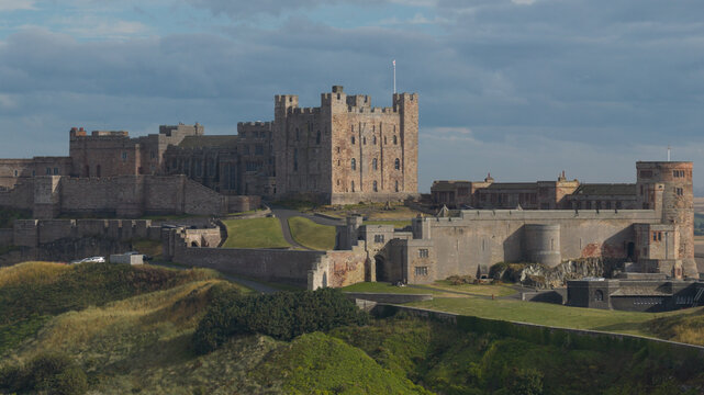 Aerial view of Bamburgh Castle with its historic stone fortifications and towers set against green grassy slopes under a cloudy sky in Bamburgh, England, United Kingdom.