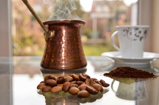 steaming copper cezve with coffee beans and ground coffee near a cup by the window