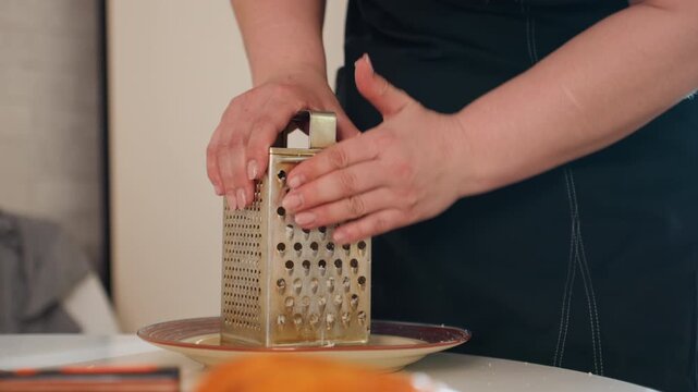 Close view of hands grating orange carrot against stainless steel box grater above shallow bowl on kitchen countertop, showing fresh food prep, texture, and steady cooking action, with hand, grating