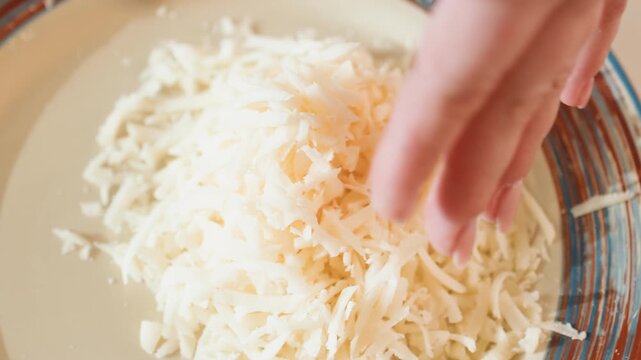 Top-down kitchen scene shows hand near rim of shallow patterned ceramic bowl filled with large mound of freshly grated white cheese, with fine shreds and loose pieces scattered around rim