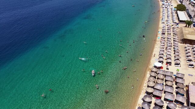 Aerial view of Toroni in Sithonia, Halkidiki, Greece, showing a busy Mediterranean beach with turquoise crystal-clear water, swimmers, sunbeds and beachfront hotels on a sunny summer day.