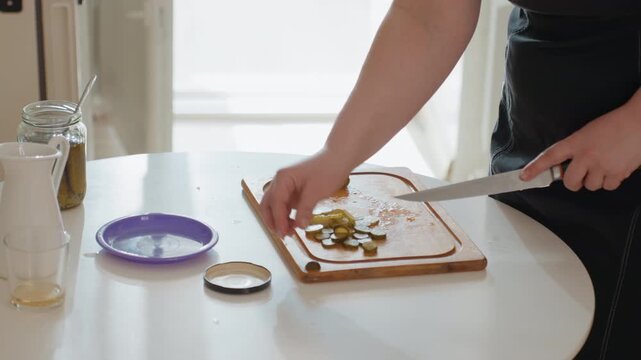 Right-handed person slices pickles with knife on wooden cutting board while left hand steadies ingredient; chopped pieces, glass jar, lid, and kitchen surface create fresh home prep scene