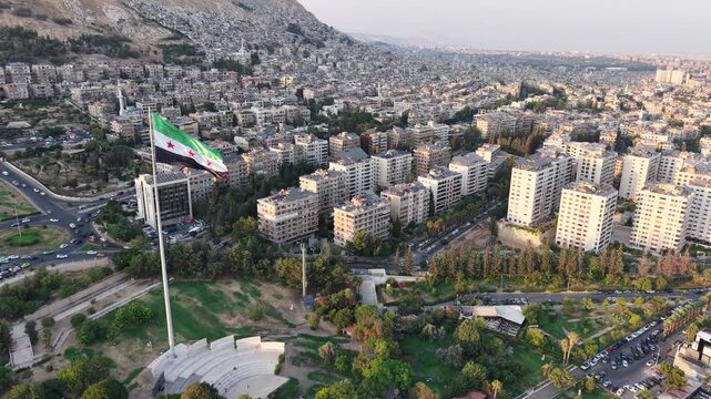 A scenic drone view shows Umayyad Square in Damascus bathed in sunset light as a massive Syrian flag waves over the landmark plaza and surrounding city landscape.