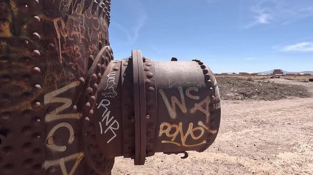Rusty steam engine boiler covered in graffiti, decaying in a desolate industrial wasteland. A forgotten, historic relic showing decay.