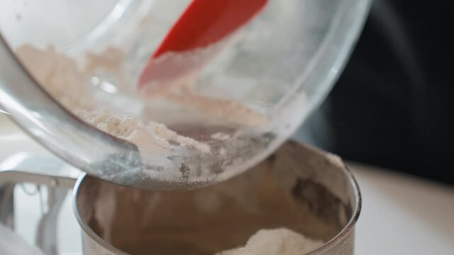 Close-up motion of ground coffee pouring from tilted clear container through metal sifter over ceramic mug, showing falling granules, soft powder cloud, warm brown texture, and steady buildup on