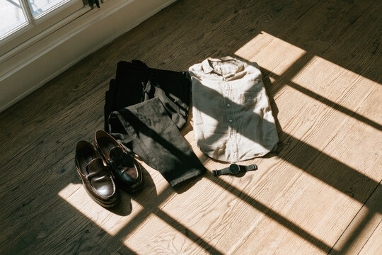 Capsule wardrobe outfit with leather loafer and wristwatch on oak floor in natural sunlight, creating organized daily preparation scene with strong window shadows and calm style