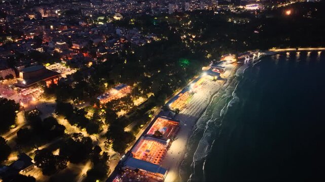 Top-down aerial night view of an illuminated seaside beach with restaurants, rows of sun loungers and umbrellas beside dark ocean waves, showing vibrant coastal nightlife and tourism.