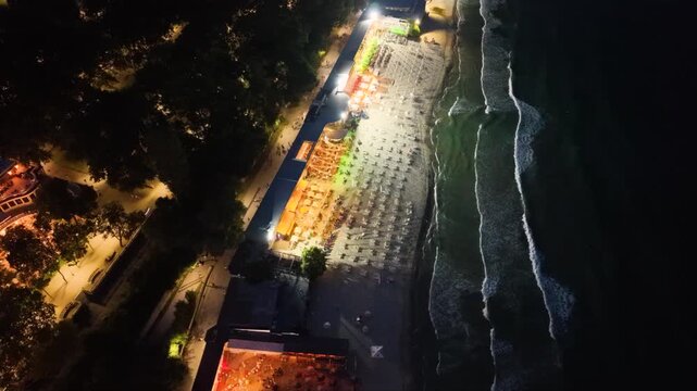 Top-down aerial night view of an illuminated seaside beach with restaurants, rows of sun loungers and umbrellas beside dark ocean waves, showing vibrant coastal nightlife and tourism.