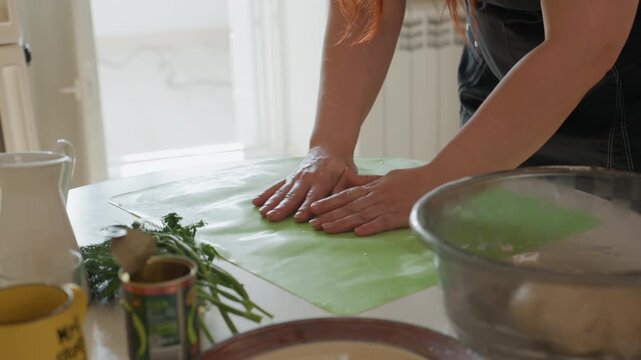 Pair of hands smoothing translucent green cloth on white table then lifting clear glass bowl to reveal round dough, with gentle pressing, folding, and warm daylight in cozy home kitchen
