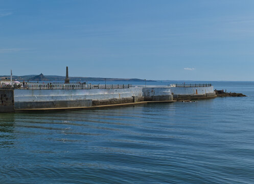 Jubilee Pool, Penzance, a curved art deco lido filled with blue seawater, under a bright sky. Distant St Michael's Mount is visible