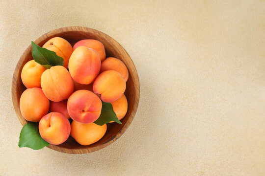 Top view of fresh ripe apricots in rustic wooden bowl with green leaves on light beige background. Healthy seasonal summer fruit, healthy food, natural nutrition, ingredient for cooking and desserts.
