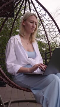 Focused blonde woman sitting in a suspended cocoon chair on a terrace, working remotely as a freelancer and typing on her laptop keyboard while enjoying the fresh air of the garden vertical video