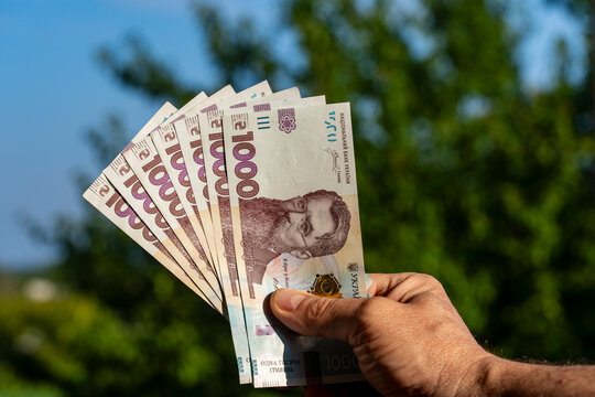 Man holding fan of Ukrainian one thousand hryvnia banknotes. Closeup of male hand holding fan of new 1000 Ukrainian hryvnia cash bills against blurry green trees and blue sky background on sunny day